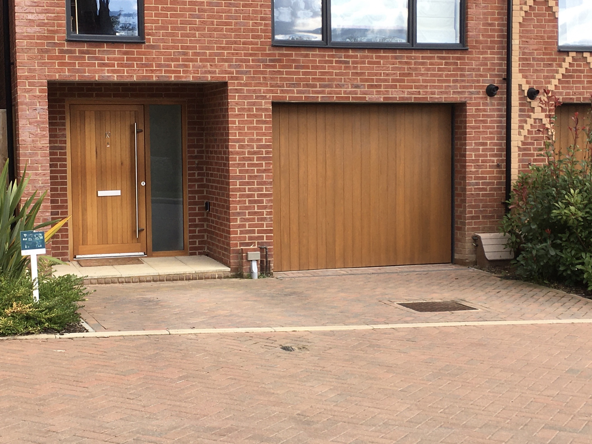 Brick townhouse facade with a wooden single garage door on the right and a matching wooden front door on the left, set within recessed brick openings and a paved driveway in front.