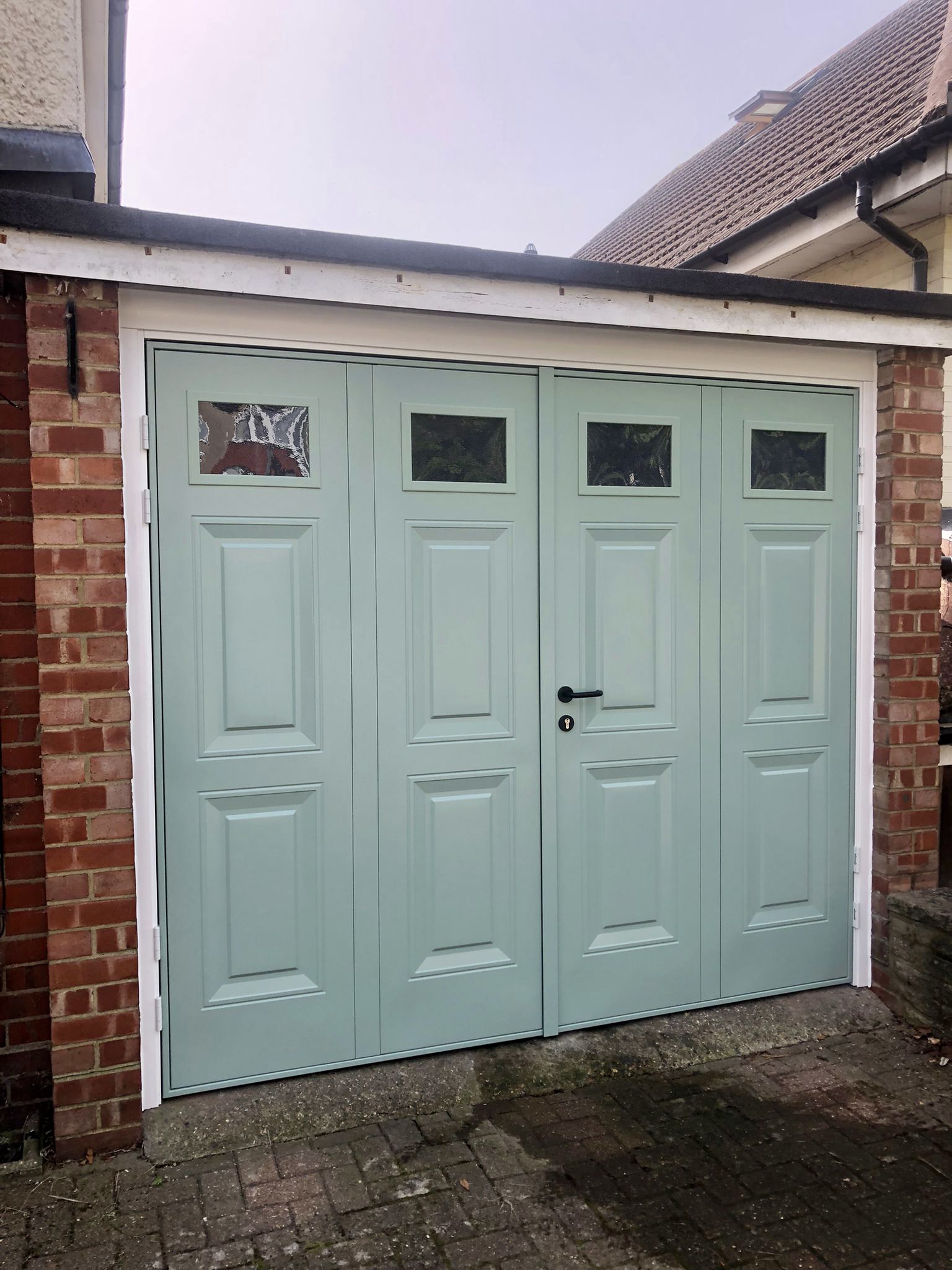 Pale green side-hinged garage doors with small glazed windows, set within a brick exterior.