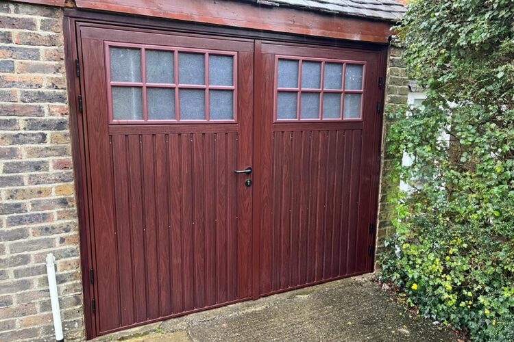 Double garage door in a dark reddish-brown wood finish with vertical panel detailing and small multi-pane windows along the top, set within a brick structure beside dense greenery.