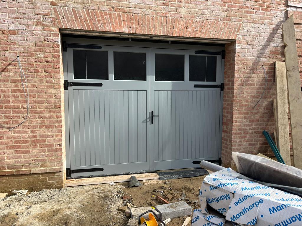 A sleek side hinged wooden garage door in grey on a brick wall